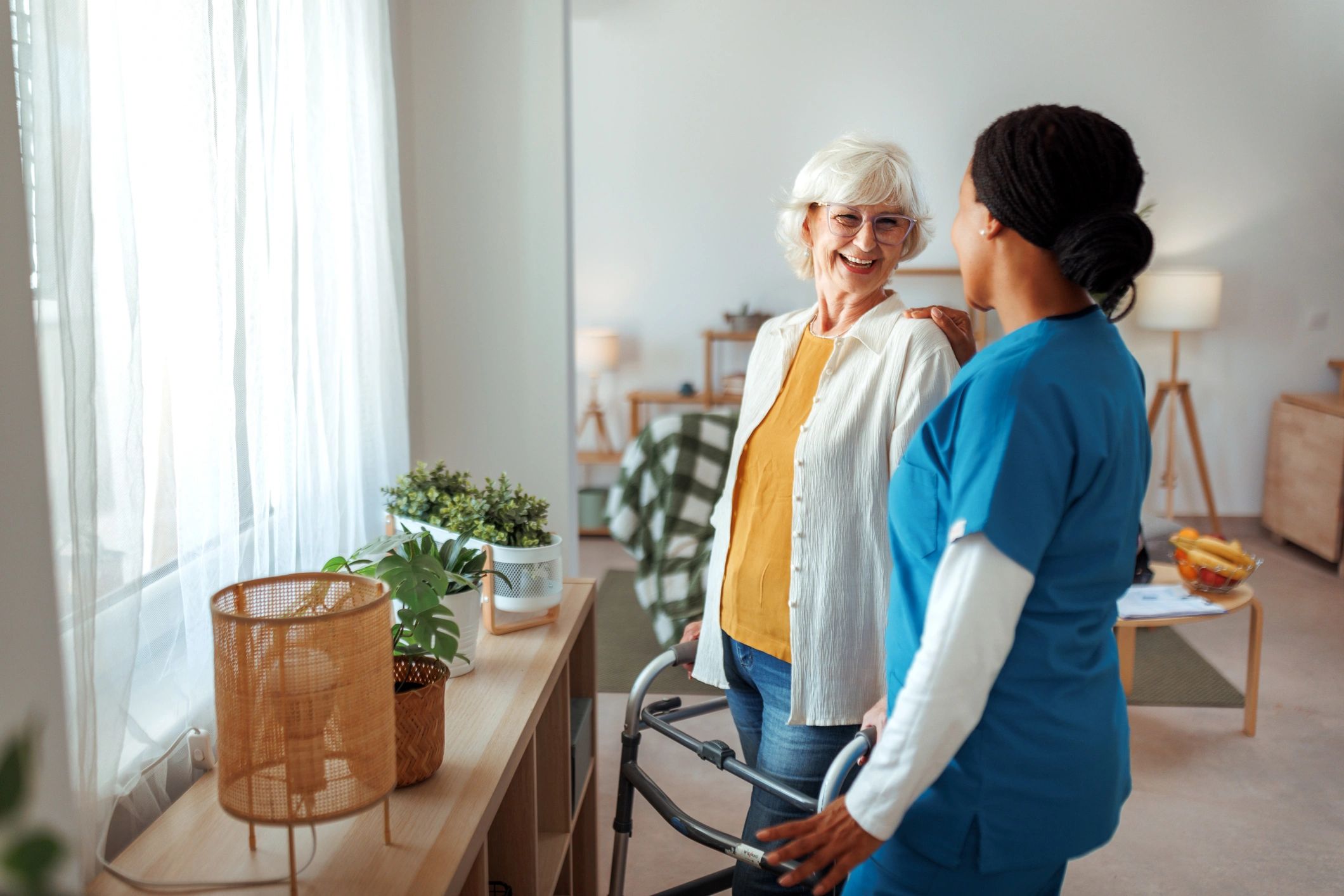 Caregiver standing with a senior woman using a walker near a window at home